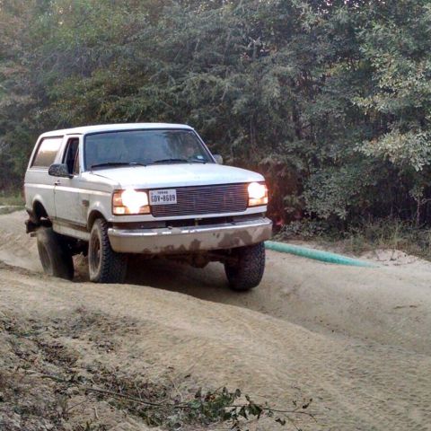 1996 Ford Bronco - A white 1996 Ford Bronco is driving off-road on a dirt path with trees in the background. The vehicle is tilted to the left as it navigates…