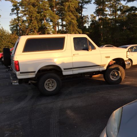 1996 Ford Bronco - A white Ford Bronco is parked in a lot with trees in the background. The vehicle is viewed from the side with the sun casting a warm glow o…
