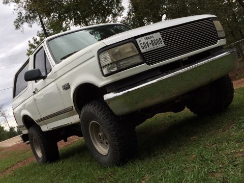 1996 Ford Bronco - A white, lifted 1990s Ford Bronco is parked on grassy terrain with trees in the background. The vehicle is viewed from a low angle, showing…