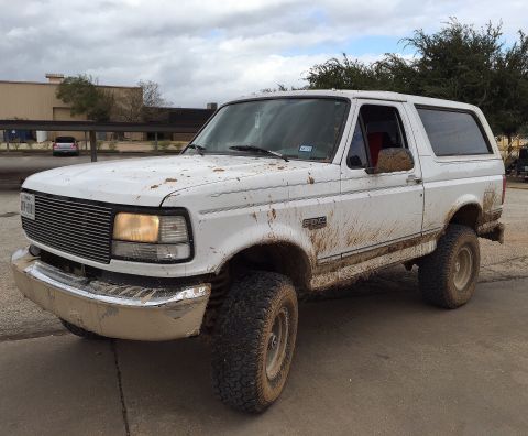 1996 Ford Bronco - A white, mud-splattered 1990s Ford Bronco is parked on a paved surface under a cloudy sky. The vehicle has a chrome grille and bumper, and…