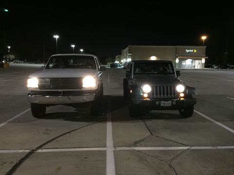 1996 Ford Bronco - Two vehicles are parked in a lot at night. On the left is a white Ford truck with its headlights on. On the right is a silver Jeep Wrangler…
