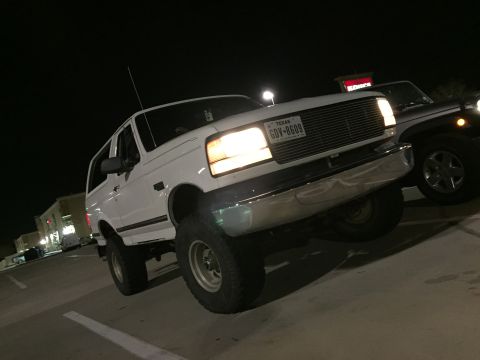 1996 Ford Bronco - A white 1990s Ford Bronco is parked in a parking lot at night, facing towards the viewer at a three-quarter front angle. The vehicle's head…