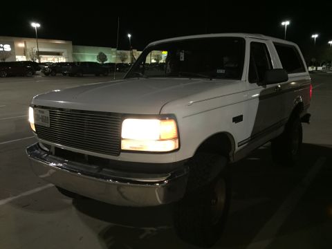 1996 Ford Bronco - A white Ford Bronco is parked in a parking lot at night, facing the camera. The vehicle's headlights are on, illuminating the front grille…