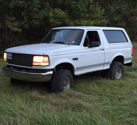 1996 Ford Bronco - A white 1990s Ford Bronco is parked in a grassy field with trees in the background. The front headlights are illuminated, and the Texas lic…