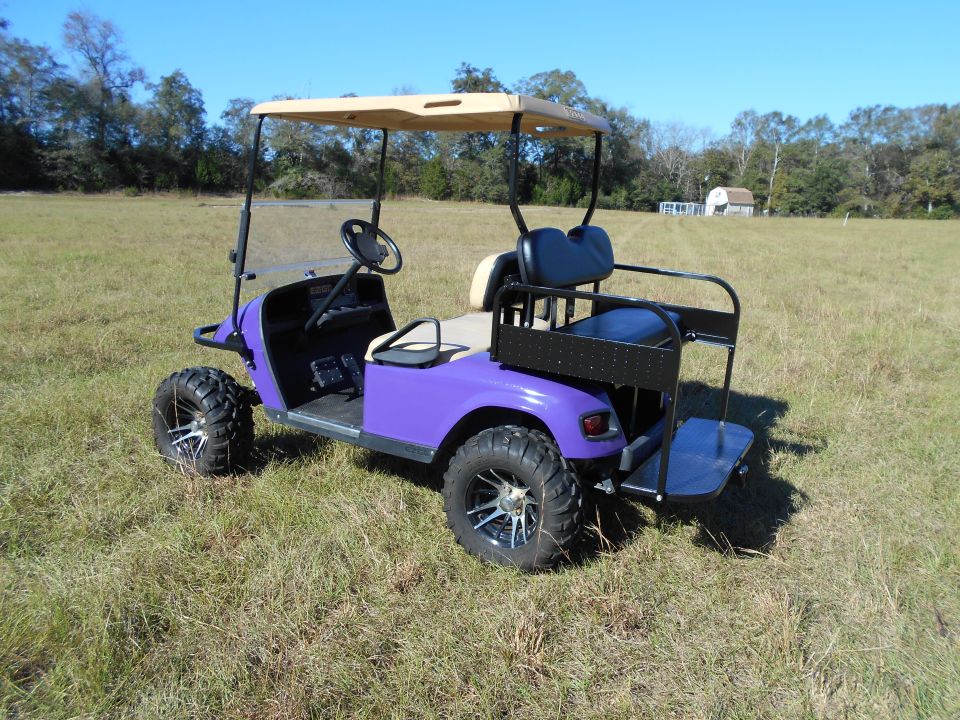 Purple golf cart with off-road tires and rear seat in a grassy field.