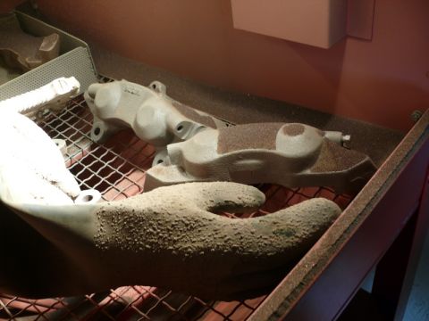 Two gray brake calipers covered in grit on a grate inside a sandblasting cabinet, with a gloved hand in the fo