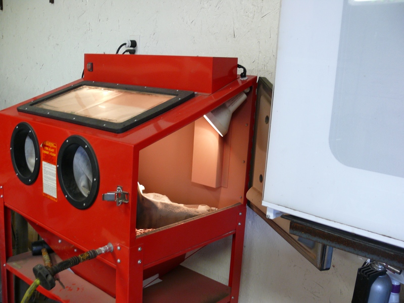 Red sandblasting cabinet with clear top and glove ports next to a white appliance.