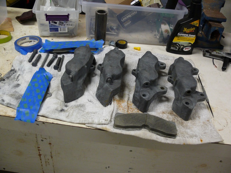 Four gray brake calipers and bolts on a workbench with blue painter's tape.