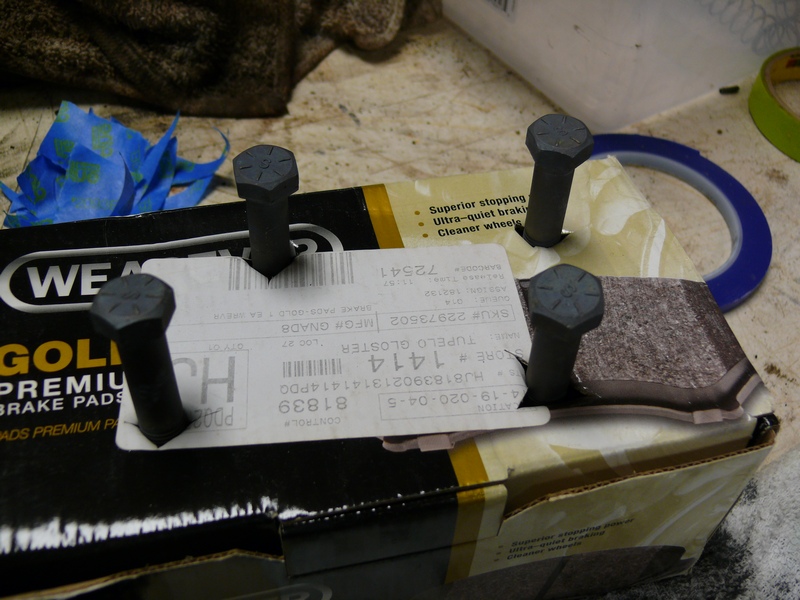 Four bolts inserted through a box of WEAREVER GOLD PREMIUM BRAKE PADS.