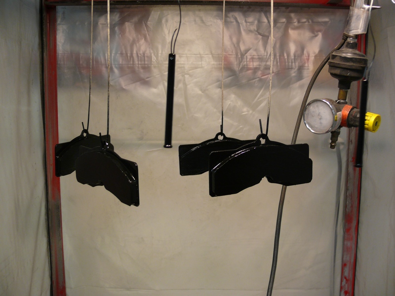 Black brake calipers hanging in a spray booth with a pressure regulator and gauge.