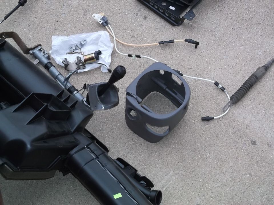 Disassembled car parts on concrete: black air duct, gear shifter, gray steering column shroud.