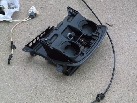 1999 Mercury Grand Marquis - A black plastic center console part with two cup holders, a cigarette lighter, and a cable attached is shown on a concrete surface. A white…