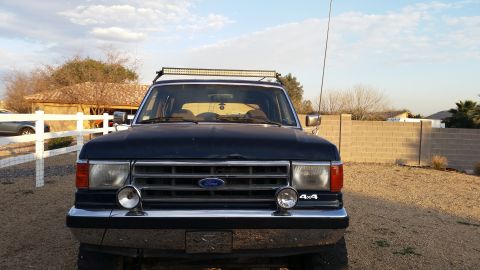 1991 Ford Bronco - Front view of a dark colored Ford Bronco parked in a gravel driveway with a white fence and a brick wall in the background. The vehicle has…