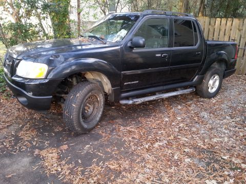 2001 Ford Explorer - A black Ford Explorer Sport Trac is parked on a gravel and leaf-covered surface in front of a wooden fence. The front passenger side of the…