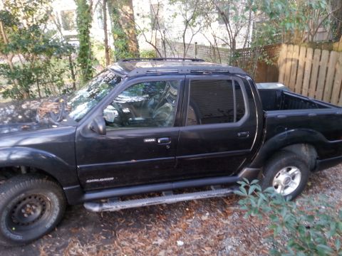 2001 Ford Explorer - The side view of a black Ford Explorer Sport Trac pickup truck is shown parked on a gravel driveway surrounded by trees and a wooden fence.…