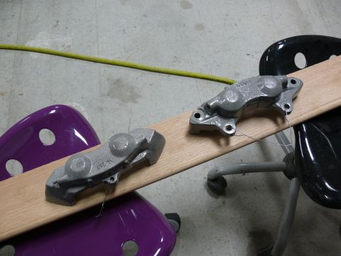 Two gray painted brake calipers suspended by wire on a wooden plank.