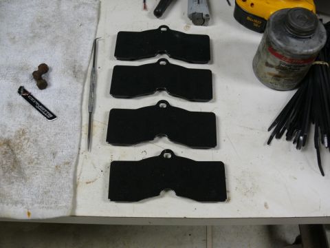 Four black brake pads, assembly lube, zip ties, Corvette sticker, and rusty metal piece on a workbench.