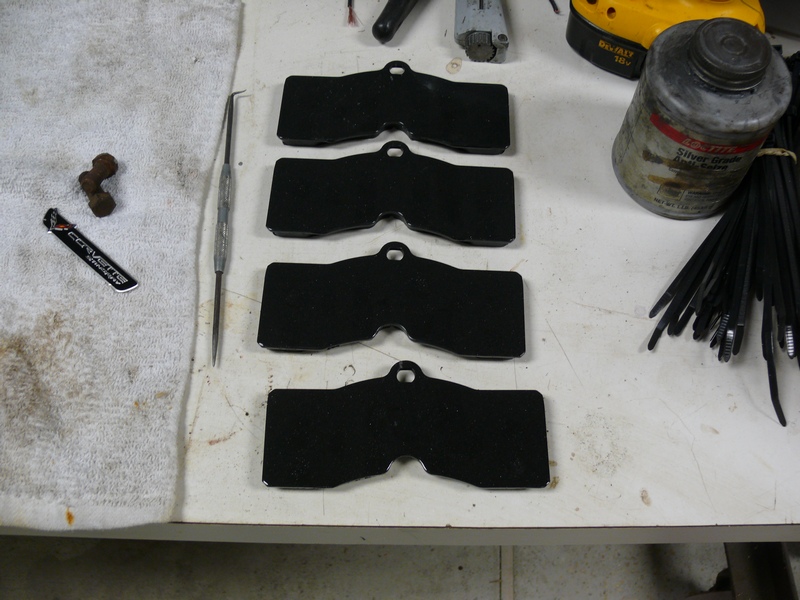 Four black brake pads, assembly lube, zip ties, Corvette sticker, and rusty metal piece on a workbench.