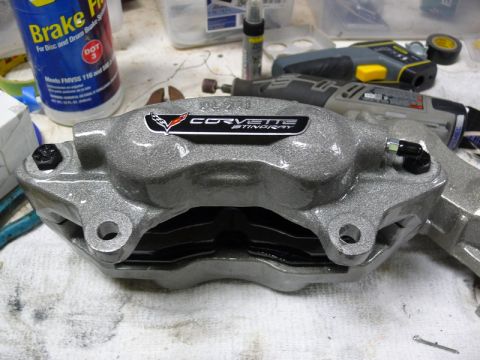 Gray Corvette Stingray brake caliper with Corvette logo, tools, and brake fluid bottle in background.