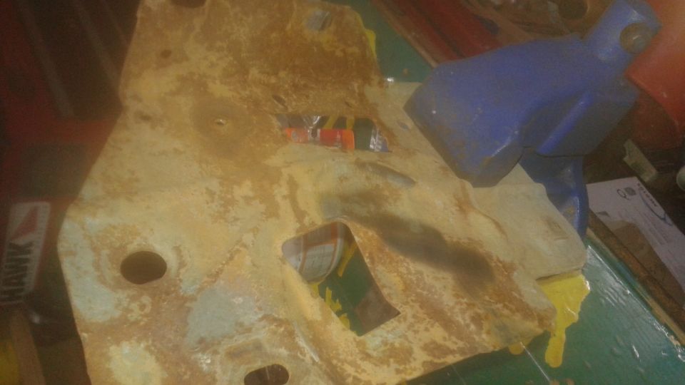 Rusty yellow metal part with cutout held in blue vise, yellow liquid dripping.