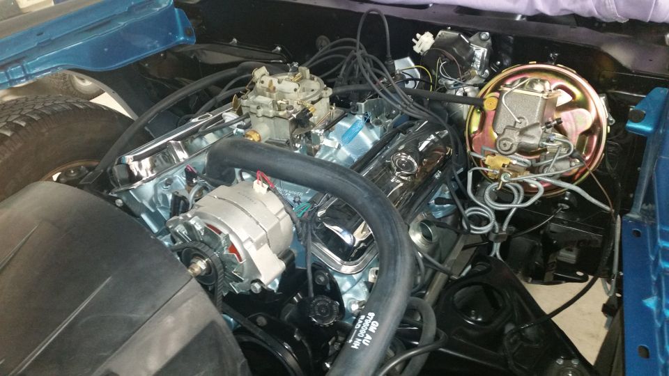 Classic car V8 engine bay with chrome valve covers, blue block, carburetor, alternator, and brake booster.