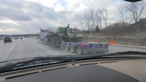 1995 Ford Bronco - A military tank is transported on a flatbed truck along a highway under a cloudy sky.