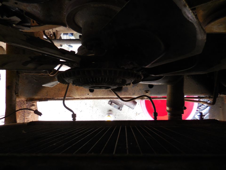 Underside of a vehicle engine bay with fan, radiator, hoses, and a red bucket.