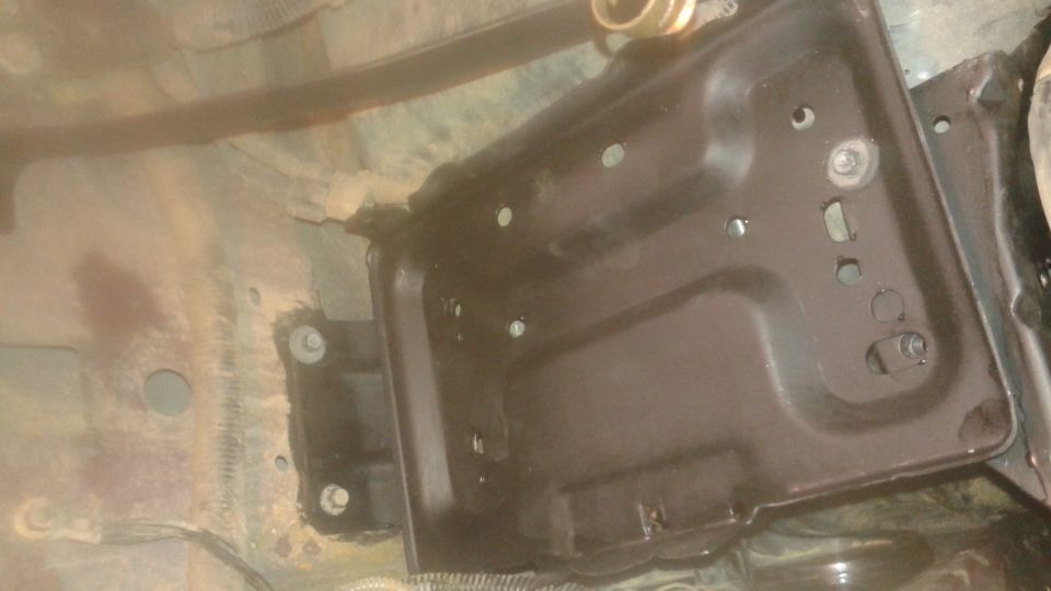 Underside view of a vehicle showing a dark metal battery tray with bolt mounts.