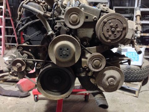 1979 Ford Bronco - A close-up, low-angle view of a heavily rusted engine assembly mounted on a red engine stand. Various pulleys, belts, and engine components…