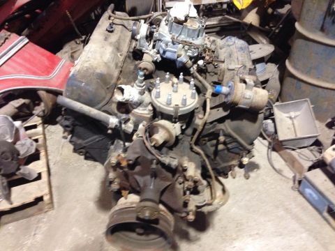1979 Ford Bronco - A dusty V8 engine is shown in a cluttered garage setting. The engine features a carburetor, distributor cap, and various hoses and wires.