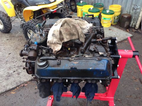 1979 Ford Bronco - An old, greasy V8 engine is mounted on a red engine stand, with a dirty rag draped over the top. Several yellow buckets and a yellow ATV ar…