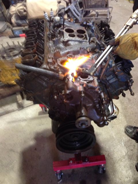 1979 Ford Bronco - A person wearing a yellow glove uses a torch to heat an engine block that is mounted on a red engine stand. The engine appears to be a V8 w…