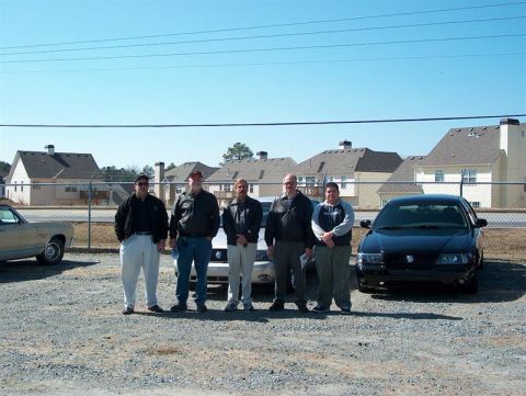 2001 Ford Crown Victoria - A group of men stand near a black sedan with a Mercury emblem, possibly a Marauder, in a parking lot.