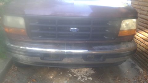 1996 Ford Bronco - The front grille and bumper of a dark red vehicle are shown. The vehicle has a chrome bumper and a black grille with a Ford emblem in the c…