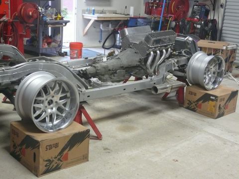 Custom car chassis with V8 engine and transmission on jack stands, with two chrome wheels on boxes.