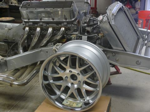 Polished aluminum engine with chrome headers on a vehicle frame, with a custom wheel in the foreground.