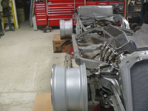 Partially assembled car chassis with V8 engine, exhaust headers, and two silver wheels in a garage.