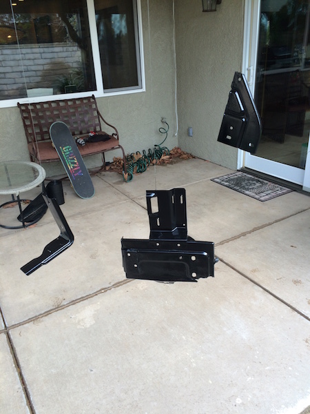 1981 Ford Bronco - Several black metal automotive parts are laid out on a concrete patio. A skateboard rests on a metal bench in the background.
