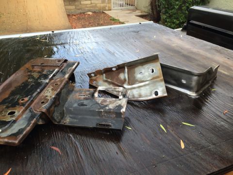 1981 Ford Bronco - Two heavily rusted metal brackets, possibly from a vehicle frame, are lying on a wet wooden surface. One bracket is larger and more complex…