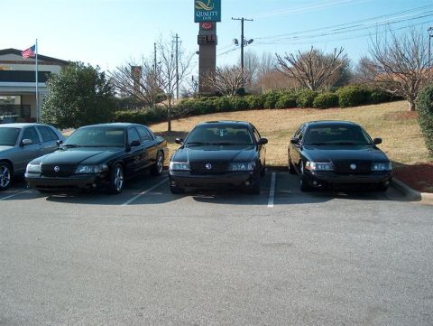2001 Ford Crown Victoria - Three black Ford Crown Victorias are parked in a lot with a silver car to the left.