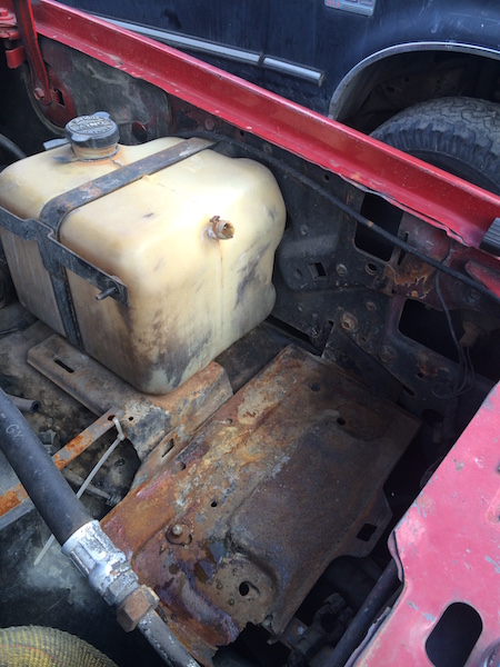 1981 Ford Bronco - The engine bay of a red vehicle shows a dirty, translucent plastic fluid reservoir secured by a rusty metal strap. A heavily rusted metal p…