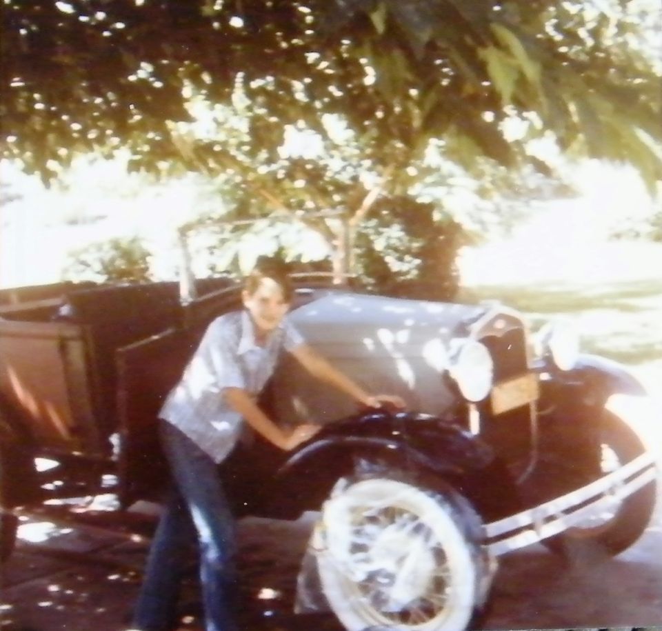 Young person next to a dark vintage convertible with a spare tire on the front wheel.