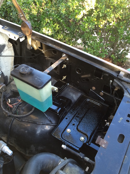 1981 Ford Bronco - The engine bay of a vehicle, showing the battery tray area which has been painted black. A windshield washer fluid reservoir with blue flui…