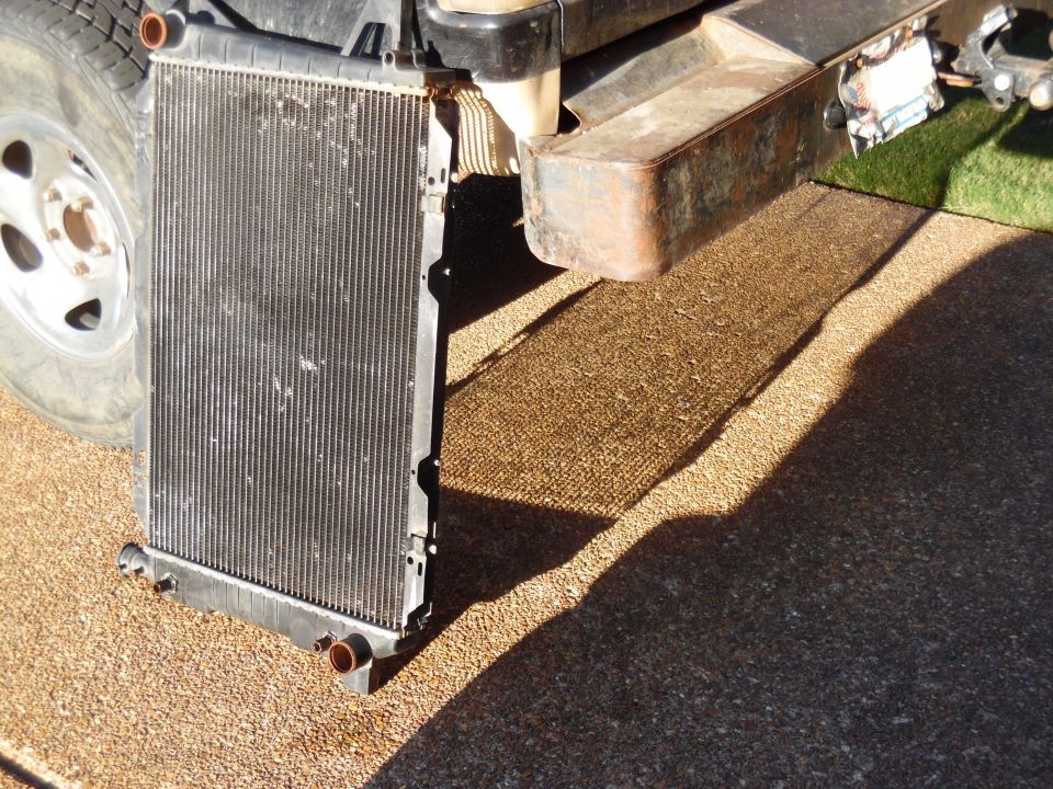 Aluminum radiator leaning against a truck frame on a pebbled surface, tire and rusty tank visible above.