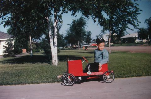 1909 Ford Model T - A young boy in a black hat sits in a red pedal car that resembles an early automobile. The pedal car has spoked wheels and a steering wheel…
