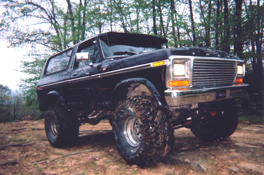 Black 1978 Ford Bronco with large off-road tires parked on dirt with trees in background