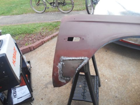 1972 Ford Bronco - Maroon front fender with a cut-out section of fresh metal repair, resting on a sawhorse in a driveway near a Hobart welder.