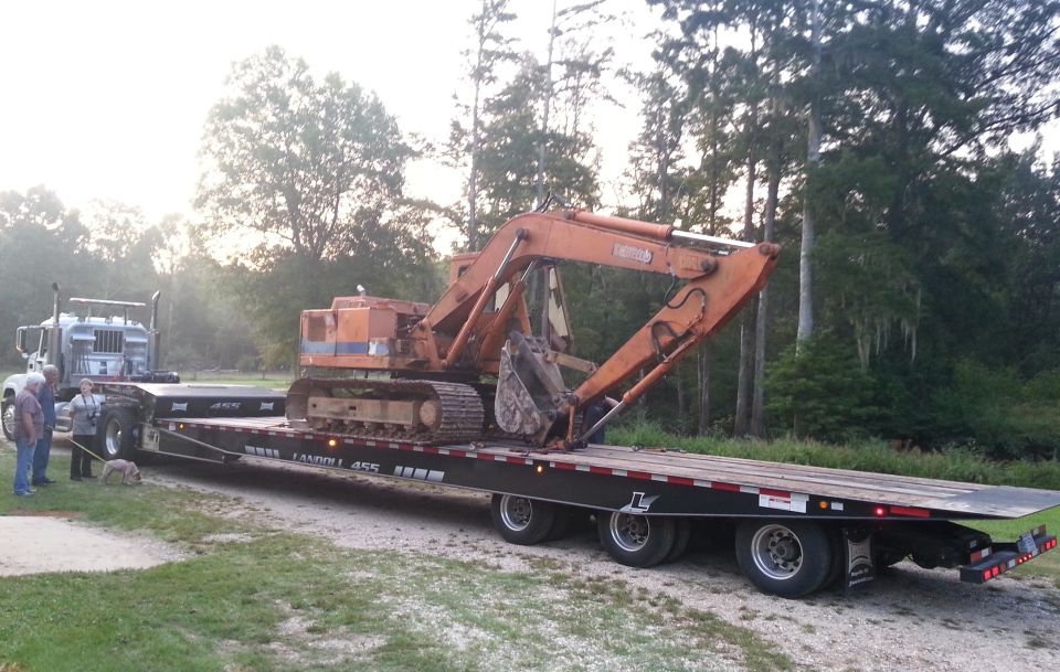 Orange excavator on a black Landoll 455 trailer hitched to a silver semi-truck, with people and a dog nearby.