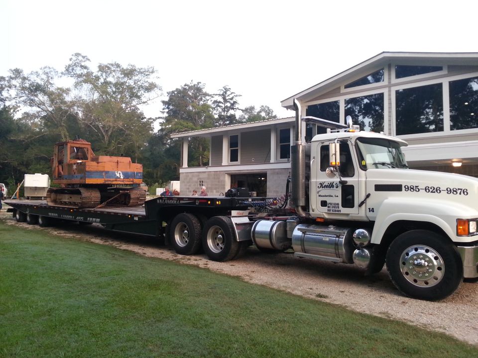 White semi-truck hauling an excavator on a flatbed trailer, parked next to a house.