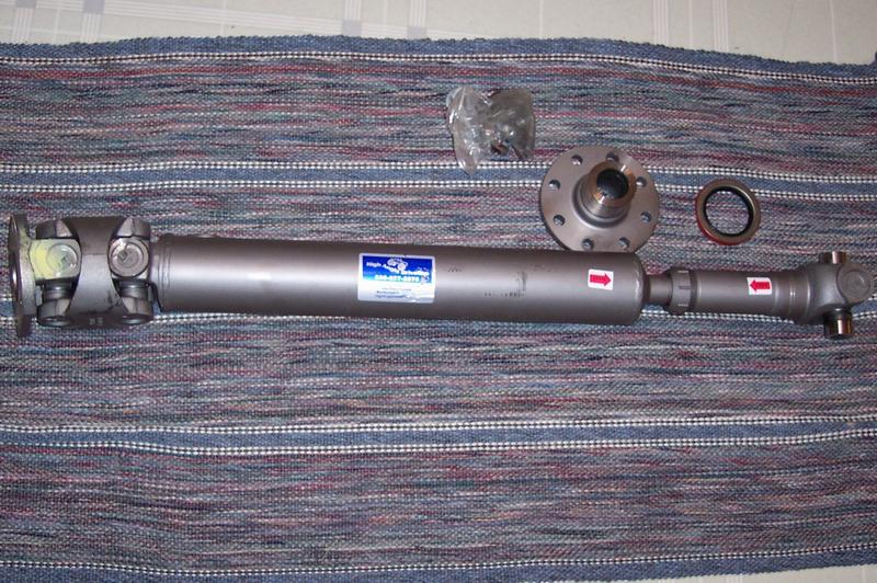 Gray driveshaft with U-joints, pinion flange, seal, and hardware bag laid on a striped rug